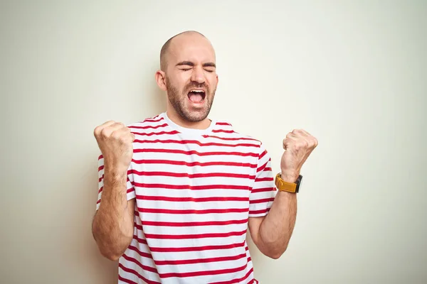 白い孤立した背景の上にカジュアルなストライプの赤いTシャツを着たあごひげを持つ若いハゲ男は 腕を上げて目を閉じて成功に驚き 驚きました 勝者のコンセプト — ストック写真