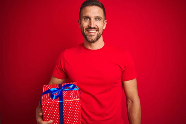 Young man holding birthday present over isolated red background with a happy face standing and smiling with a confident smile showing teeth