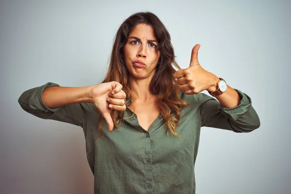 Young beautiful woman wearing green shirt standing over grey isolated background Doing thumbs up and down, disagreement and agreement expression. Crazy conflict