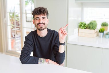 Kameraya bakarak yan el ve parmak ile işaret giyen sıradan kazak yüzünde kocaman bir gülümseme ile beyaz masaya oturan genç adam.