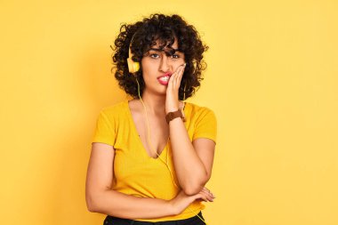 Kıvırcık saçlı Arap kadın izole sarı arka plan üzerinde kulaklık kullanarak müzik dinlerken yorgun görünümlü ve çapraz kolları ile depresyon sorunları ile sıkılmış düşünme.