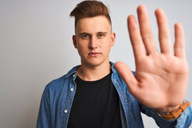 Denim gömlek giyen genç yakışıklı adam ciddi ve kendinden emin ifade, savunma jesti ile dur işareti yapıyor açık el ile izole beyaz arka plan üzerinde duran