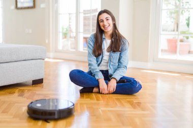 Yerleri temizlemek için otomatik süpürge kullanan genç bir kadın makine ev işi robotu kontrol ediyor.