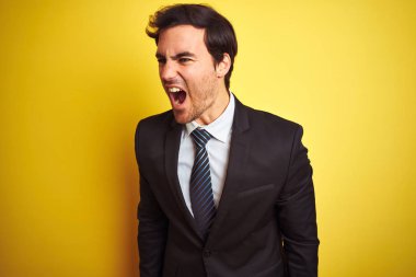 Genç yakışıklı işadamı giyen takım elbise ve kravat ayakta izole sarı arka plan öfkeli ve deli çığlık sinirli ve öfkeli, öfke ile bağırarak. Öfke ve agresif kavram.