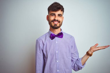Dövme mor gömlek ve yay kravat giyen genç adam izole beyaz arka plan gülümseyen neşeli sunan ve kamera bakarak el avuç ile işaret.