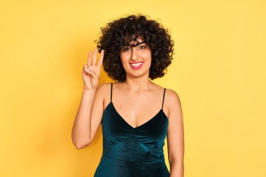 İzole sarı arka plan üzerinde zarif bir elbise giyen kıvırcık saçlı genç arap kadın gösteren ve emin ve mutlu gülümserken parmaksayısı üç işaret.