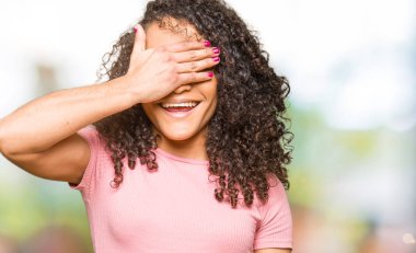 Genç güzel kadın gülümsüyor ve gözleri için sürpriz kapsayan yüz el ile gülüyor pembe t-shirt giyen kıvırcık saçlı. Kör kavramı.