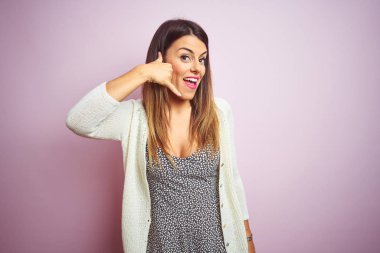 Genç güzel kadın pembe izole arka plan üzerinde duran telefonda konuşurken gibi el ve parmakları ile telefon jest yapıyor gülümseyerek. İletişim kavramları.