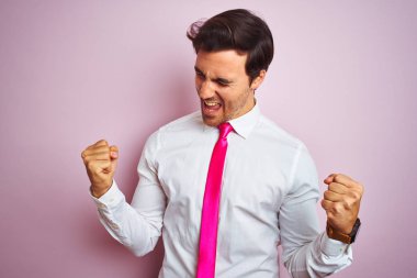 Genç yakışıklı işadamı gömlek giyen ve kravat izole pembe arka planda ayakta çok mutlu ve heyecanlı silah ile kazanan jest yaparak, gülümseyen ve başarı için çığlık kaldırdı. Kutlama konsepti.