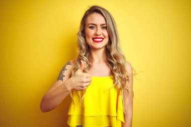 Sarı izole arka planda ayakta t-shirt giyen genç attactive kadın el ile jest kadar mutlu başparmak yapıyor. Başarı gösteren kameraya bakarak ifadeyi onaylama.