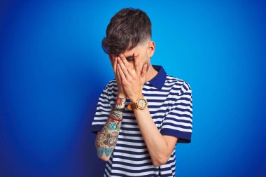 Dövmeli genç adam ağlıyor iken elleri ile yüz kapsayan üzücü ifade ile izole mavi arka plan üzerine çizgili polo ayakta giyiyor. Depresyon konsepti.