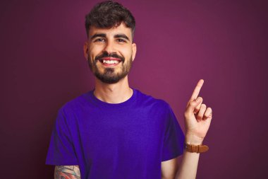 Yüz üzerinde büyük bir gülümseme ile izole mor arka planda ayakta tişört giyen dövme ile genç adam, kamera bakarak yan el ve parmak ile işaret.