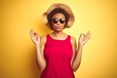 Sarı izole arka plan üzerinde yaz şapka ve güneş gözlüğü giyen Afrikalı Amerikan kadın Relax ve gözleri ile gülümseyen meditasyon jest parmaklar ile kapalı. Yoga konsepti.
