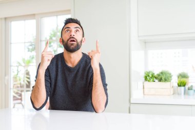 Evde rahat kazak giyen yakışıklı İspanyol adam hayran ve bakmak ve parmaklar ve yükseltilmiş kollar ile işaret şaşırttı.