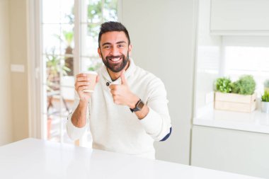 Yakışıklı İspanyol adam bir kağıt fincan mutlu büyük gülümseme ile ok işareti, parmak yukarı başparmak, mükemmel işareti yapıyor bir kahve içme