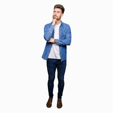 Çene düşünme hakkında soru, dalgın ifade üzerine el ile rahat denim ceket giyen genç yakışıklı sarışın adam. Düşünceli yüzüyle gülümsüyor. Şüphe kavramı.