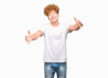 Afro saç ile genç yakışıklı adam rahat beyaz t-shirt kucaklamak için açık kollar ile gülümseyen kamera bakıyor giyiyor. Mutluluğu kucaklayan neşeli ifade.