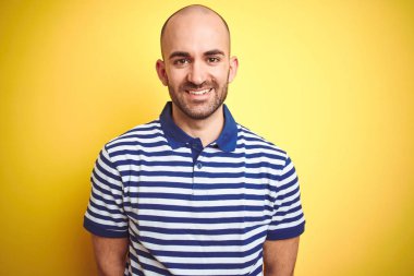 Yüz üzerinde mutlu ve serin bir gülümseme ile sarı izole arka plan üzerinde rahat çizgili mavi t-shirt giyen sakal ile genç kel adam. Şanslı kişi.