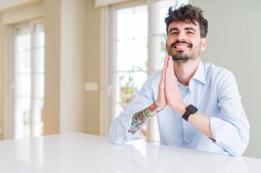 Genç Business adam oturan üzerinde beyaz tablo dua ile eller birlikte yalvararak için af gülümseyen emin.