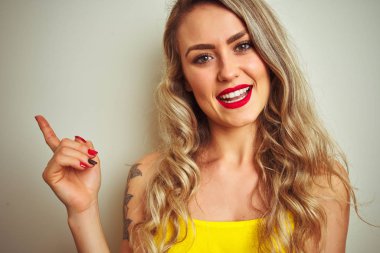 Beyaz izole arka plan üzerinde ayakta sarı t-shirt giyen genç güzel kadın çok mutlu yan el ve parmak ile işaret