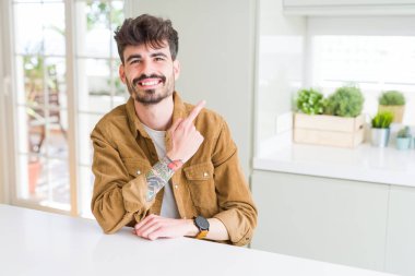 Beyaz masada neşeli oturan rahat ceket giyen genç adam yüzünde mutlu ve doğal ifade ile yan yana el ve parmak ile işaret eden yüz bir gülümseme ile neşeli
