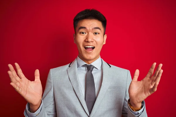 Asian chinese businessman wearing grey jacket and tie standing over isolated red background celebrating crazy and amazed for success with arms raised and open eyes screaming excited. Winner concept