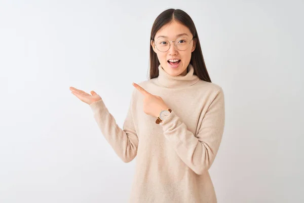 孤立した白い背景の上にタートルネックのセーターとメガネを身に着けている若い中国人女性は 手を差し伸べ 指で指を向けながら カメラに驚き 微笑んでいます — ストック写真