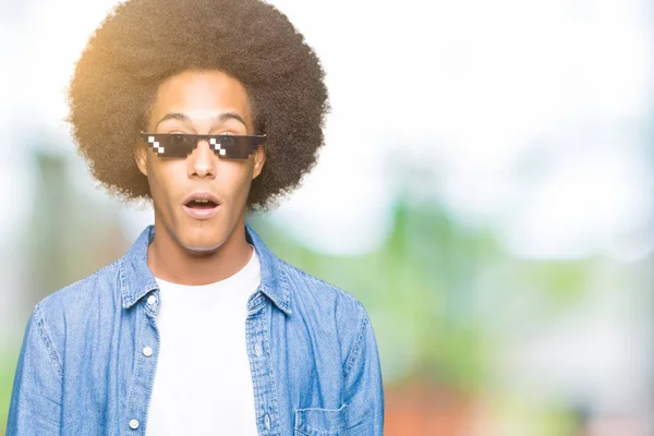 Young african american man with afro hair wearing thug life glasses afraid and shocked with surprise expression, fear and excited face.