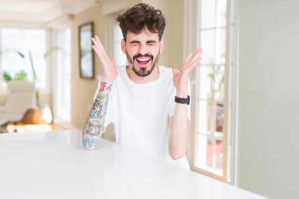 Hombre Joven Con Camiseta Casual Sentado Mesa Blanca Celebrando Loco —  Fotos de Stock