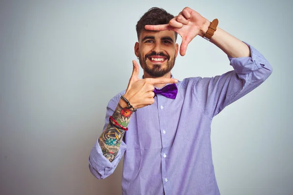 Young man with tattoo wearing purple shirt and bow tie over isolated white background smiling making frame with hands and fingers with happy face. Creativity and photography concept.