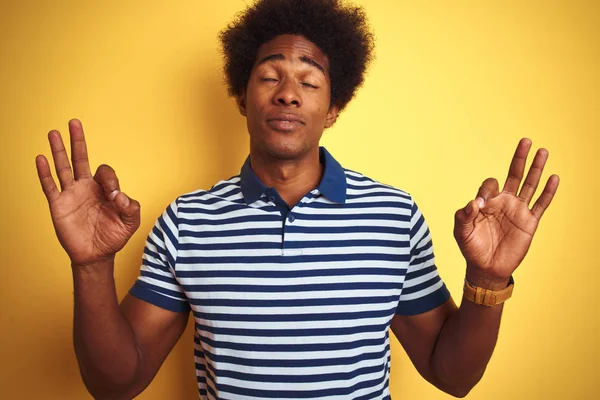 American man with afro hair wearing navy striped polo standing over isolated yellow background relax and smiling with eyes closed doing meditation gesture with fingers. Yoga concept.