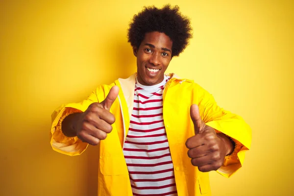African american man with afro hair wearing rain coat standing over isolated yellow background approving doing positive gesture with hand, thumbs up smiling and happy for success. Winner gesture.