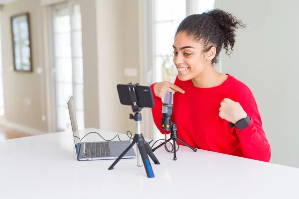 Young african american woman doing video call using smartphone camera and microphone looking confident with smile on face, pointing oneself with fingers proud and happy.