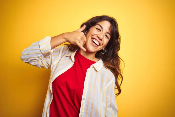 赤いTシャツとストライプのシャツを着た若い美しい女性は 電話で話すような手と指で電話ジェスチャーをして微笑む黄色の孤立した背景の上に 概念の伝達 — ストック写真