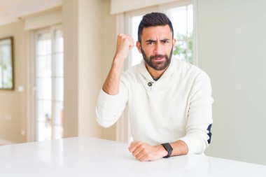 Yakışıklı İspanyol adam evde rahat beyaz kazak giyiyor öfkeli ve deli yükseltme yumruk sinirli ve öfkeli öfke ile bağırırken. Öfke ve agresif kavram.