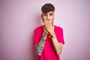 Dövme ile genç adam izole pembe arka plan üzerinde duran t-shirt ayakta hata elleri ile ağız kapsayan şok. Gizli konsept.