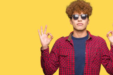 Kapalı giyen güneş gözlüğü rahatla afro saçlı yakışıklı delikanlı ve gülümseyen gözlerle parmak ile meditasyon jest yapıyor. Yoga kavramı.