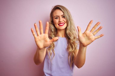 Pembe izole arka planda duran mor t-shirt ayakta giyen genç güzel kadın ve emin ve mutlu gülümseyen iken parmak numarası on ile işaret.