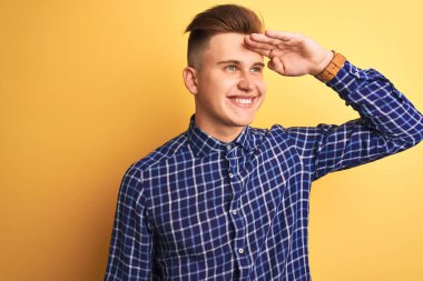 Genç yakışıklı adam izole sarı arka plan üzerinde duran rahat gömlek giyen çok mutlu ve başının üzerinde el ile uzak görünümlü gülümsüyor. Arama konsepti.