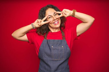 Kırmızı izole arka plan üzerinde önlük üniforma giyen orta yaş yaşlı kadın yüz üzerinde parmakları ile barış sembolü yapıyor, zafer gösteren neşeli gülümseyen