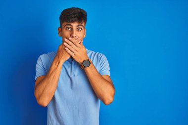 Izole mavi arka plan üzerinde duran rahat polo giyen genç hintli adam hata için elleri ile ağız kapsayan şok. Gizli kavram.