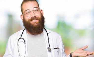 Sakal giyen tıbbi ceket ile genç sarışın doktor adam her iki el açık avuç gösteren gülümseyen, sunma ve reklam karşılaştırma ve denge