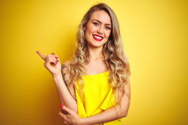 Yüze büyük bir gülümseme ile sarı izole arka plan üzerinde ayakta t-shirt giyen genç attactive kadın, kamera bakarak yan el ve parmak ile işaret.