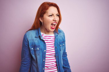 Güzel kızıl saçlı kadın kot gömlek giyiyor ve çizgili t-shirt üzerinde izole pembe arka plan öfkeli ve deli çığlık sinirli ve öfkeli, öfke ile bağırıyor. Öfke ve agresif kavram.