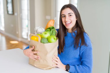 Evde bakkal dolu bir kağıt çanta tutan gülümseyen genç güzel kadın