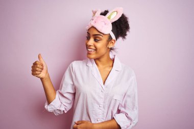 Genç Afrikalı Amerikalı kadın pijama ve izole pembe arka plan üzerinde Maske giyen gururlu görünümlü, yan hareket başparmak yukarı yapıyor gülümseyen