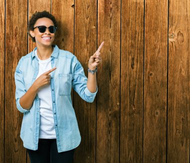 Güzel genç Afro-Amerikan kadın gülümseyerek ve iki elleri ve parmakları ile tarafına işaret kamera bakarak izole arka plan üzerinde güneş gözlüğü takıyor.