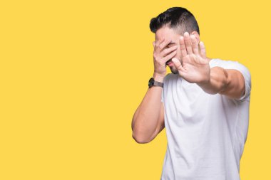 Genç adam gözleri elleriyle kapsayan izole arka plan üzerinde rahat beyaz t-shirt giyen ve stop jest ile acı ve korku ifadesi. Utandım ve negatif kavramı.