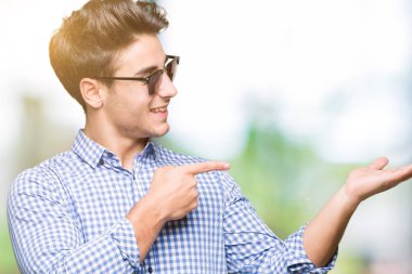 Yakışıklı delikanlı hayrete ve el ile sunulması ve parmak ile işaret ederken kameraya gülümseyen izole arka plan üzerinde güneş gözlüğü takıyor.