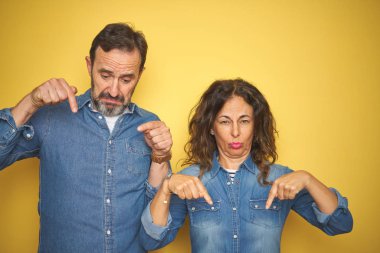 Güzel orta yaş çift birlikte izole sarı arka plan üzerinde duran üzgün ve üzgün görünümlü, parmakları ile yön gösteren, mutsuz ve depresif.